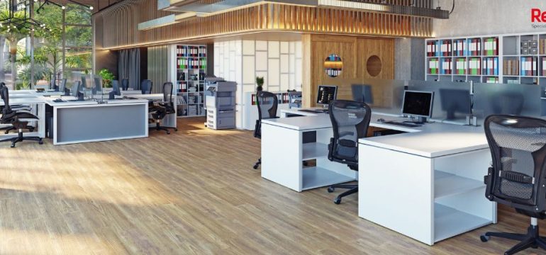 Modern open-plan office with several empty desks and computer monitors, wooden floors, large windows letting in natural light, and shelves with books and files in the background.