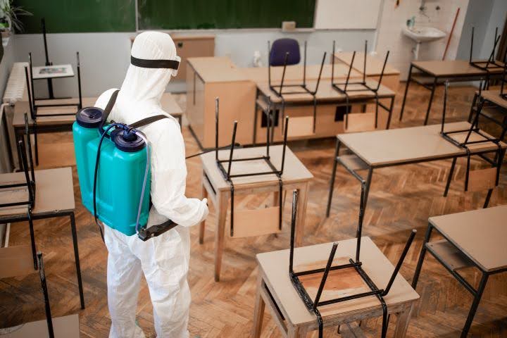 A specialist hygiene technician in full protective gear, spraying disinfectant inside a classroom to protect against the transmission of infectious pathogens