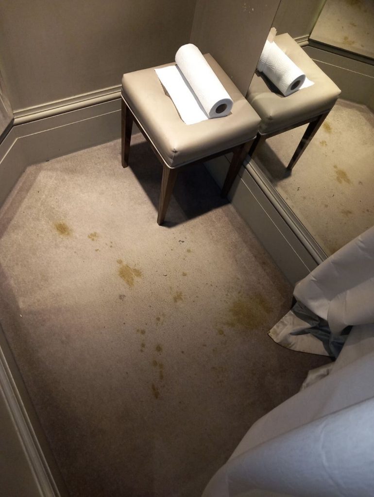 Yellow and brown bio hazard stains splattered across a beige carpeted floor next to a leather stool and mirror in a fitting or dressing room.