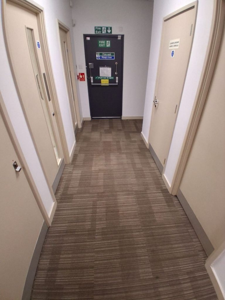A hallway with multiple beige doors, featuring professionally cleaned brown linear-pattern carpet tiles, with all previous stains removed, leading toward a dark green fire exit door with green signage.