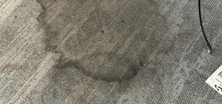 Large, dark circular liquid stain on a grey textured carpet tile floor in a commercial office.