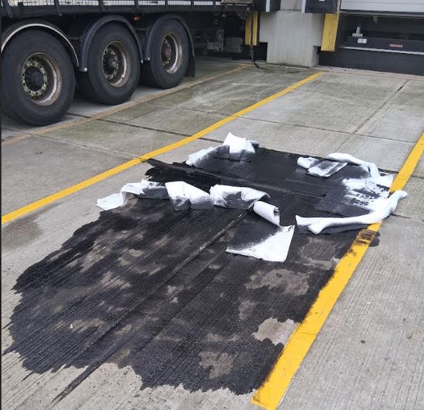 Large black oil spill on concrete outside a loading dock, partially covered with white absorbent pads and spill socks next to a semi-truck.