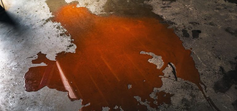Large puddle of dark orange automotive fluid or oil spilled across a grimy concrete garage floor.