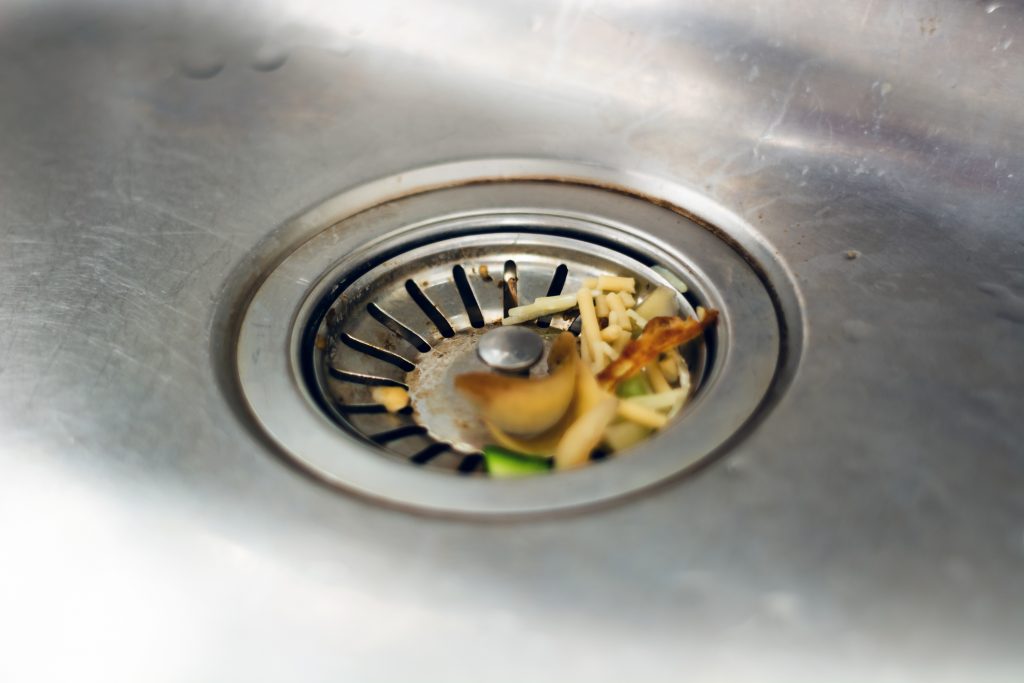 A close-up of a kitchen sink drain with food scraps, including vegetable peels and shredded pieces, caught in the metal strainer.