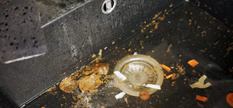 A close-up of a kitchen sink filled with dirty water, vegetable scraps, and food debris. The drain is partially visible, and the black sink surface is wet and grimy.
